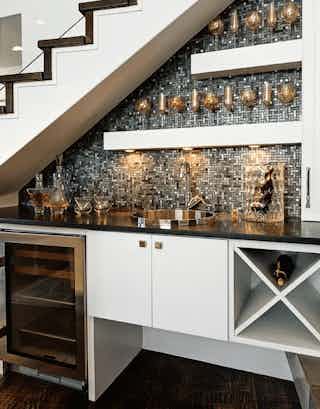 A wet bar installed underneath a white staircase that has a black tile backsplash