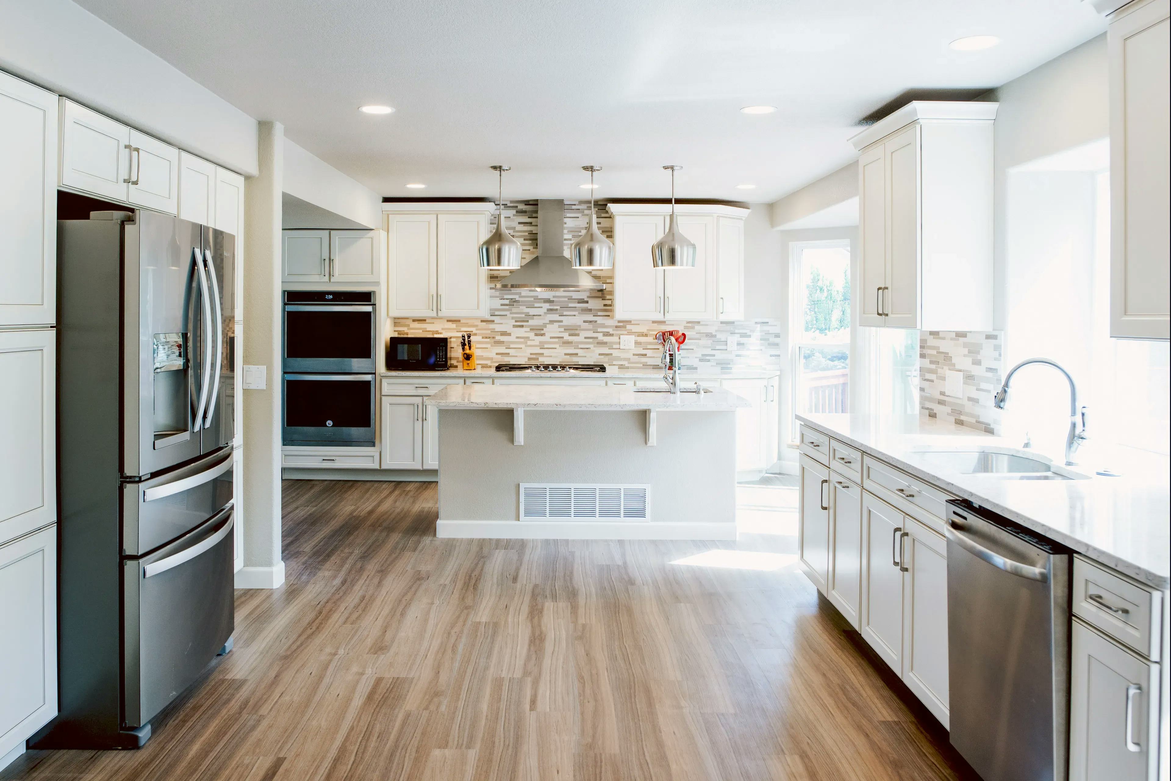 A wide angle photo showing an entirely new remodeled minimal kitchen with white cabinets, countertops, wooden floors and stainless steel appliances
