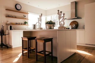 A photograph of a new kitchen remodel with a modern / minimal feel, square stools, white cabinets, a spacious center island and wooden shelves with white countertops 