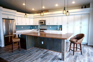 A remodeled kitchen with a blue tiled splash wall, wooden floors, a hammered copper sink, white cabinets, and a wooden breakfast bar and custom hanging lighting